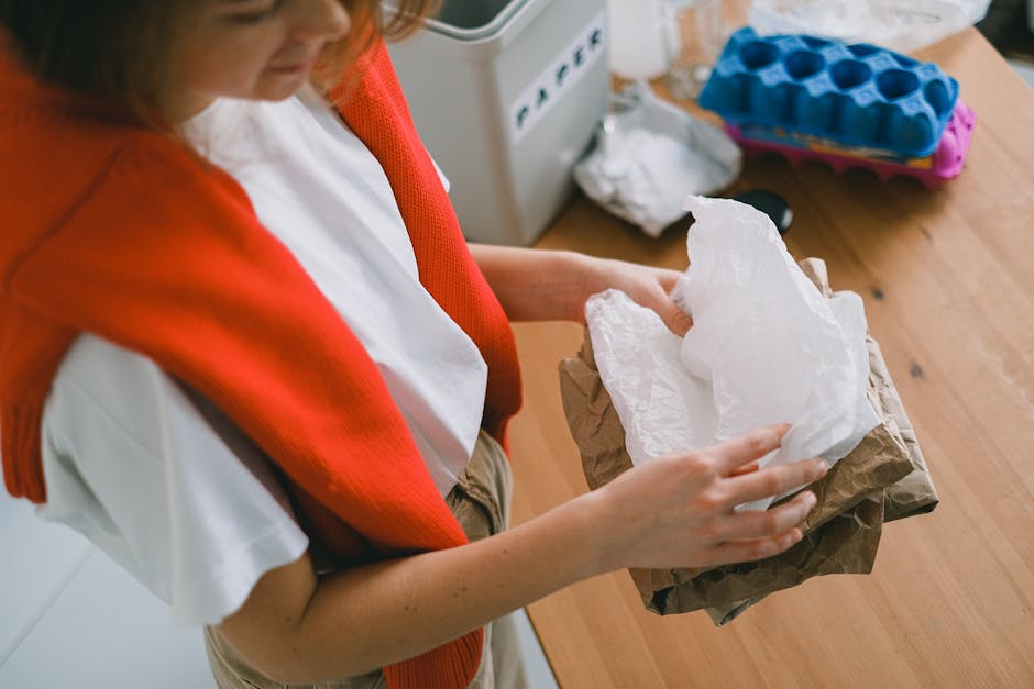 A person with light skin, wearing a white shirt and a red knitted vest, is holding a stack of crumpled white and brown paper wrapping or packaging materials, with their hands visible in the image. The