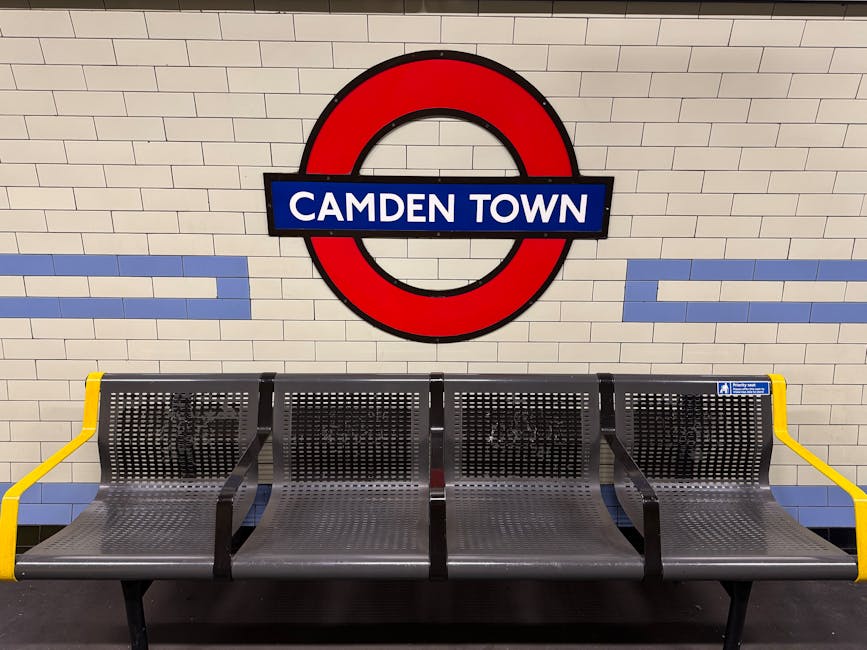 A row of four black metal benches with perforated seats and backrests, positioned in front of a tiled wall at Camden Town underground station. The benches have yellow armrests on both ends, providing